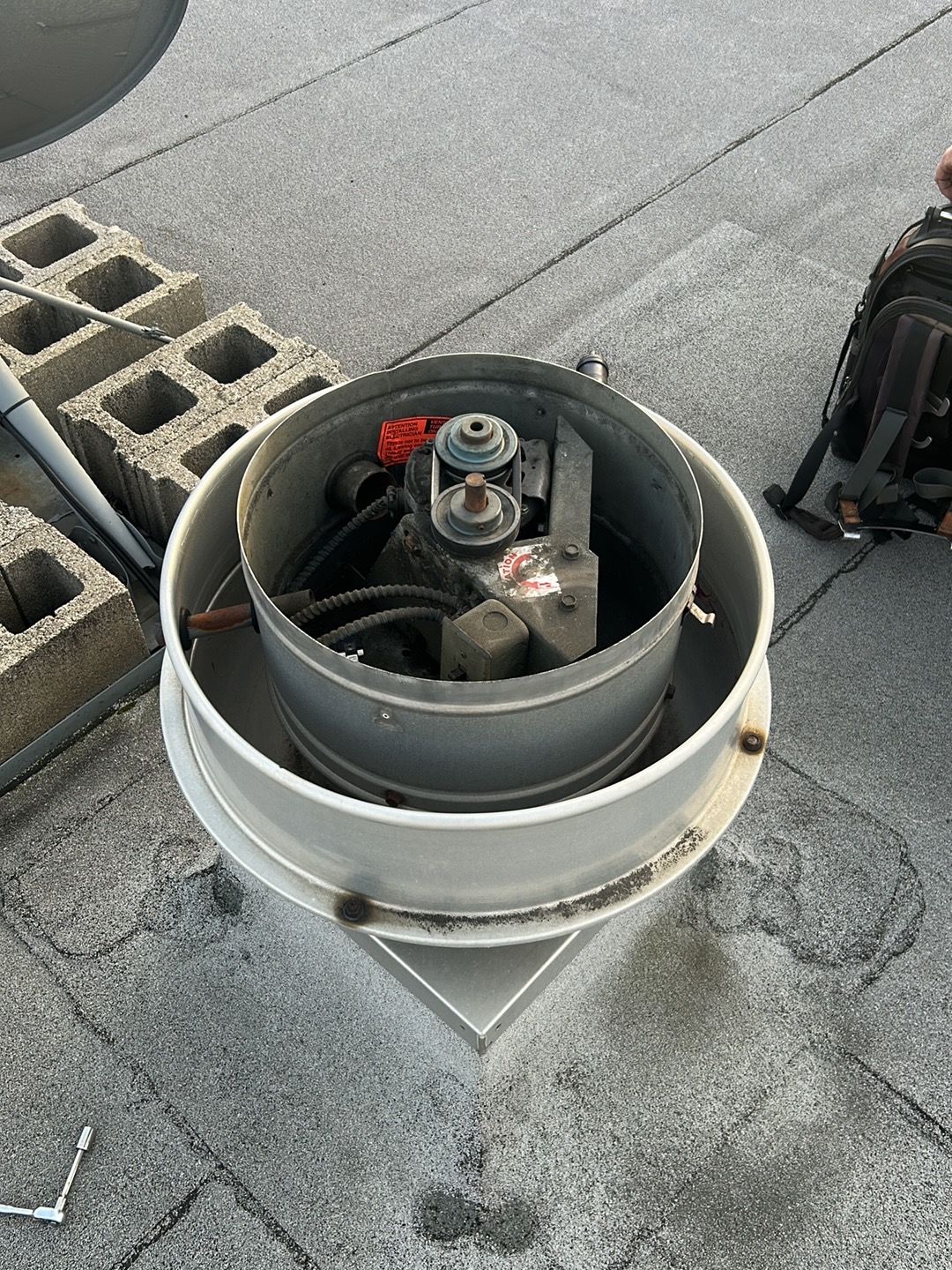 An open industrial roof exhaust fan with a belt-driven motor mechanism sitting on a flat rooftop near a concrete block.