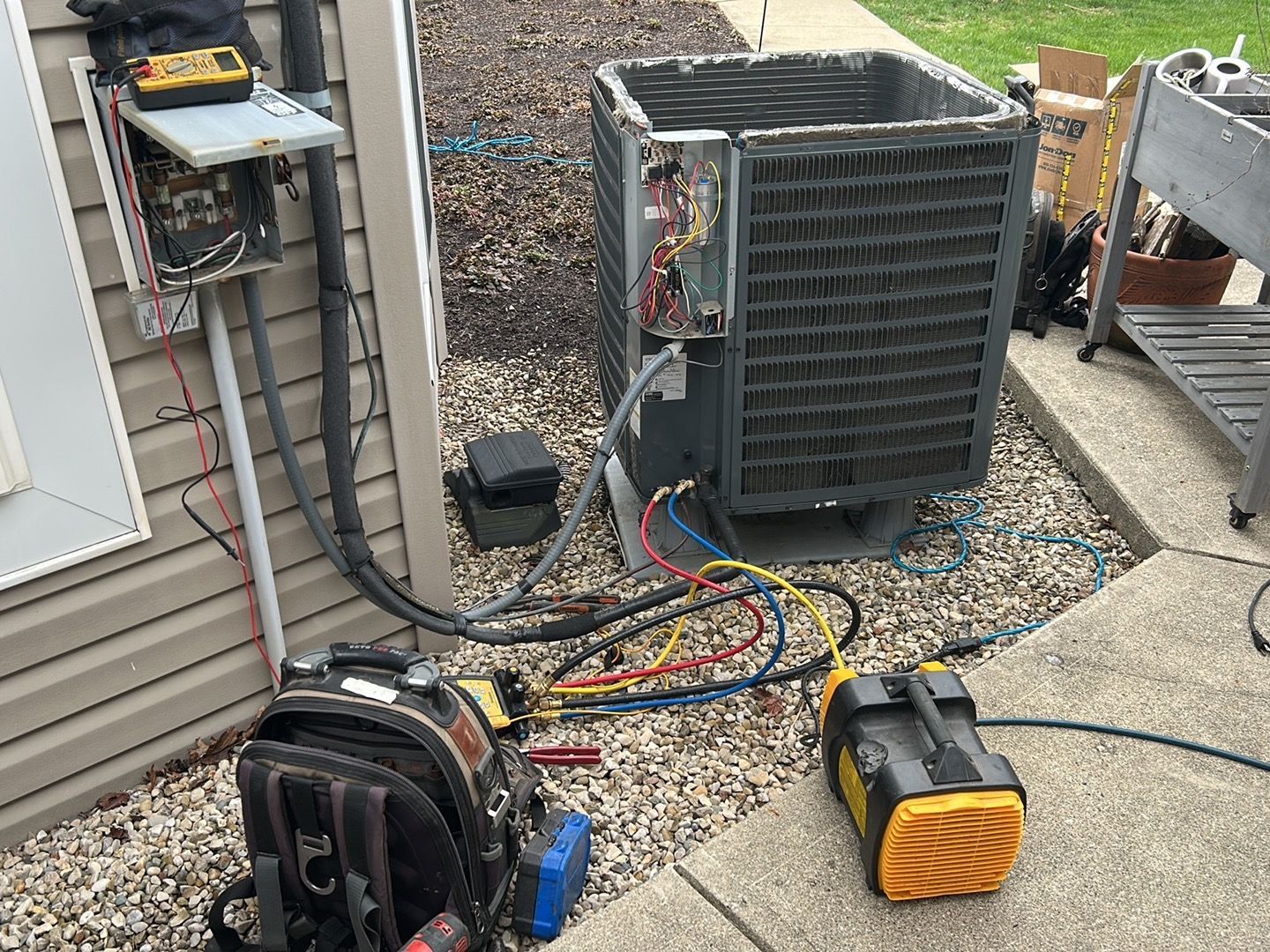An outdoor air conditioning unit with its service panel removed, connected to refrigerant gauges and a vacuum pump.