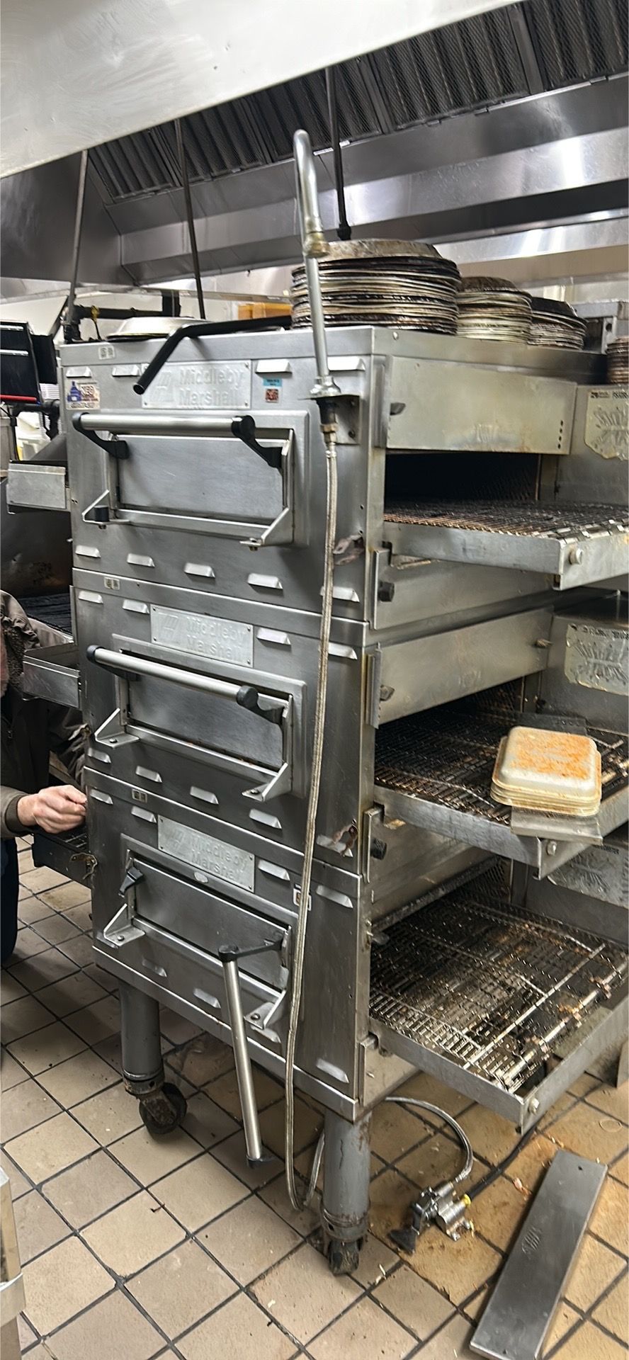 A multi-tiered commercial conveyor belt oven inside a kitchen with tiled flooring and a ventilation hood above.