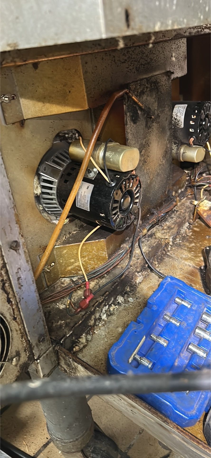 Close-up of a furnace burner assembly with a black motor, brass components, and a blue tool case on a dirty floor.