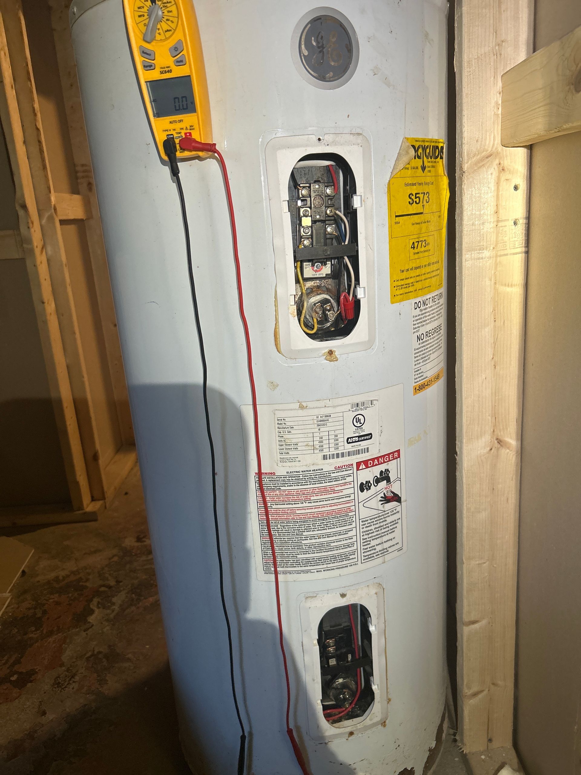 A multimeter tests an electric water heater with exposed upper and lower access panels in a residential space.