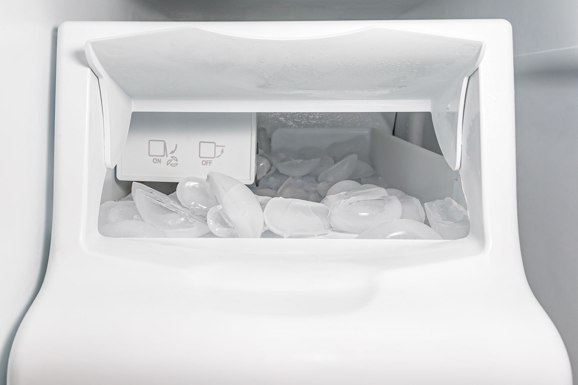 An ice dispenser inside a refrigerator freezer, showing a bin filled with ice cubes and a white control switch.