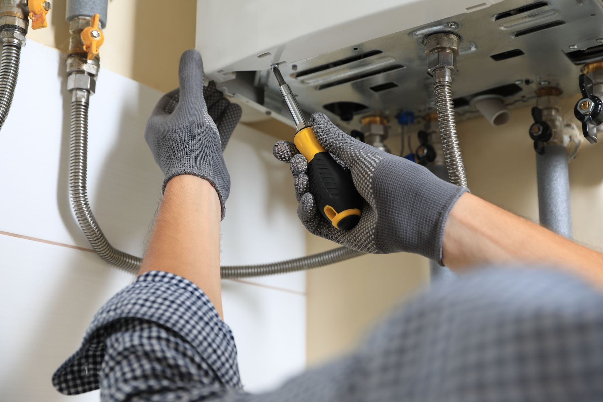 A person in gray work gloves using a screwdriver to repair a white wall-mounted boiler with exposed metal pipes.