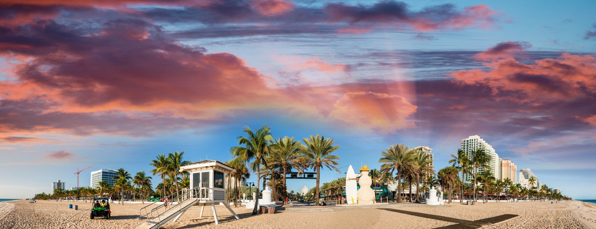 Sunset beach resort with palm trees, white buildings, and dramatic pink-orange clouds
