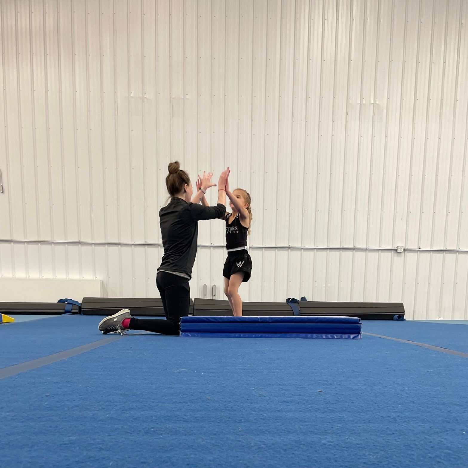 Two girls are kneeling on a blue mat in a gym.
