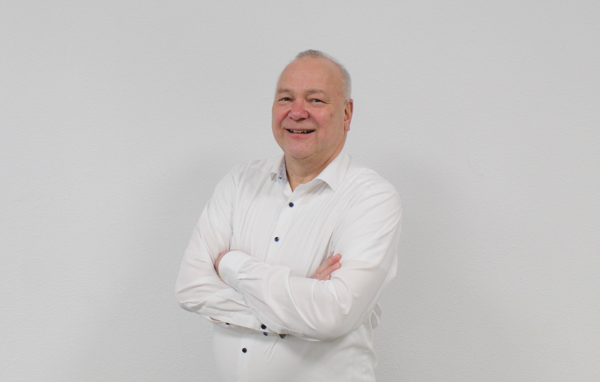 Een man in een wit overhemd staat met zijn armen over elkaar en lacht.