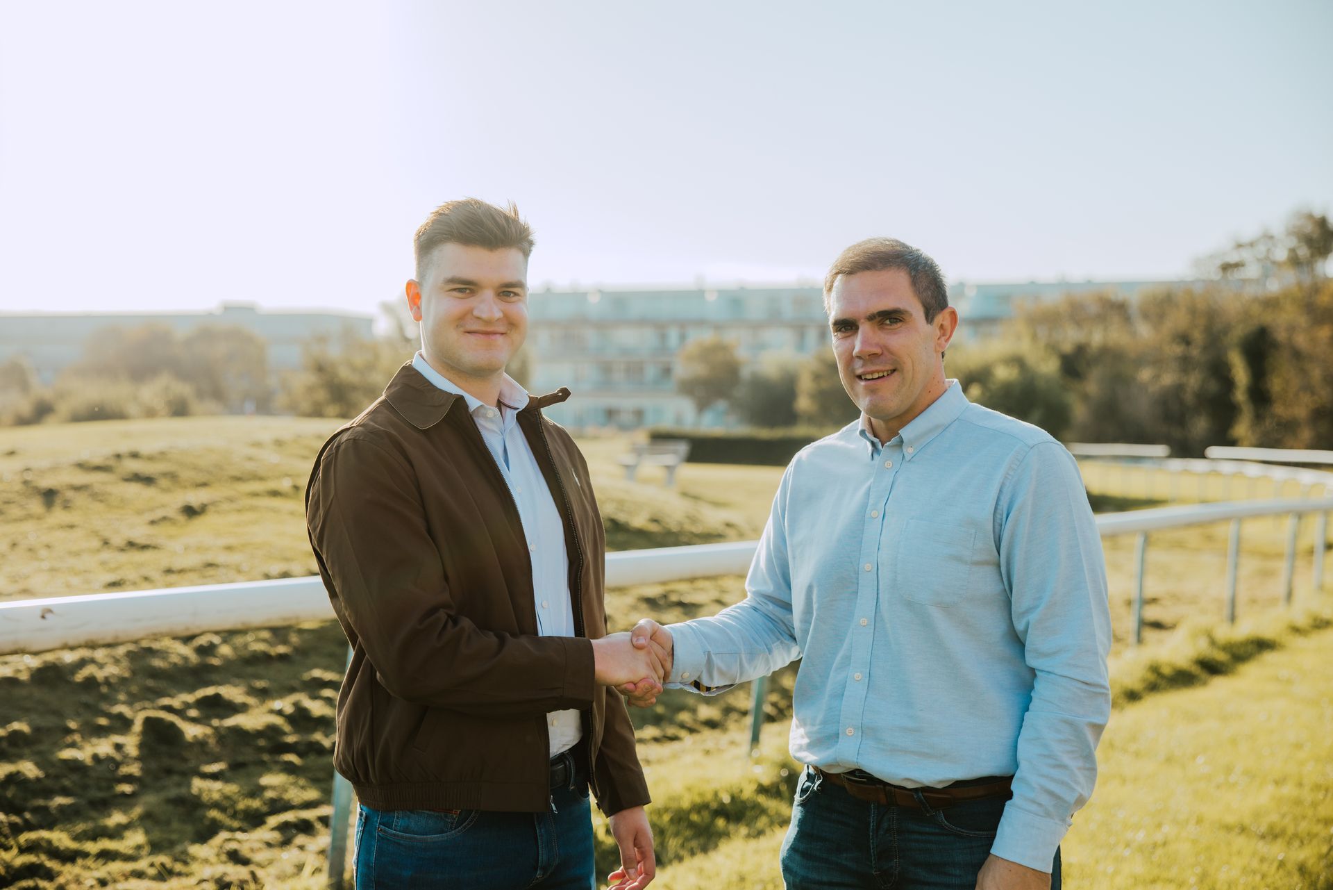 Twee mannen schudden elkaar buiten de hand, waarschijnlijk een deal. Groen gras, zonnig, gebouwen op de achtergrond.