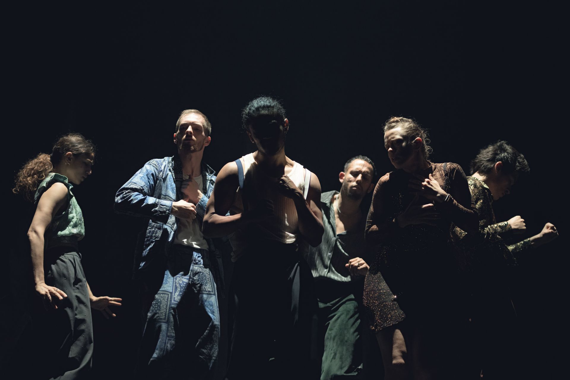 Dancers in dark clothing on a stage. They are illuminated by spotlights, some are gesturing, and one is wearing denim.