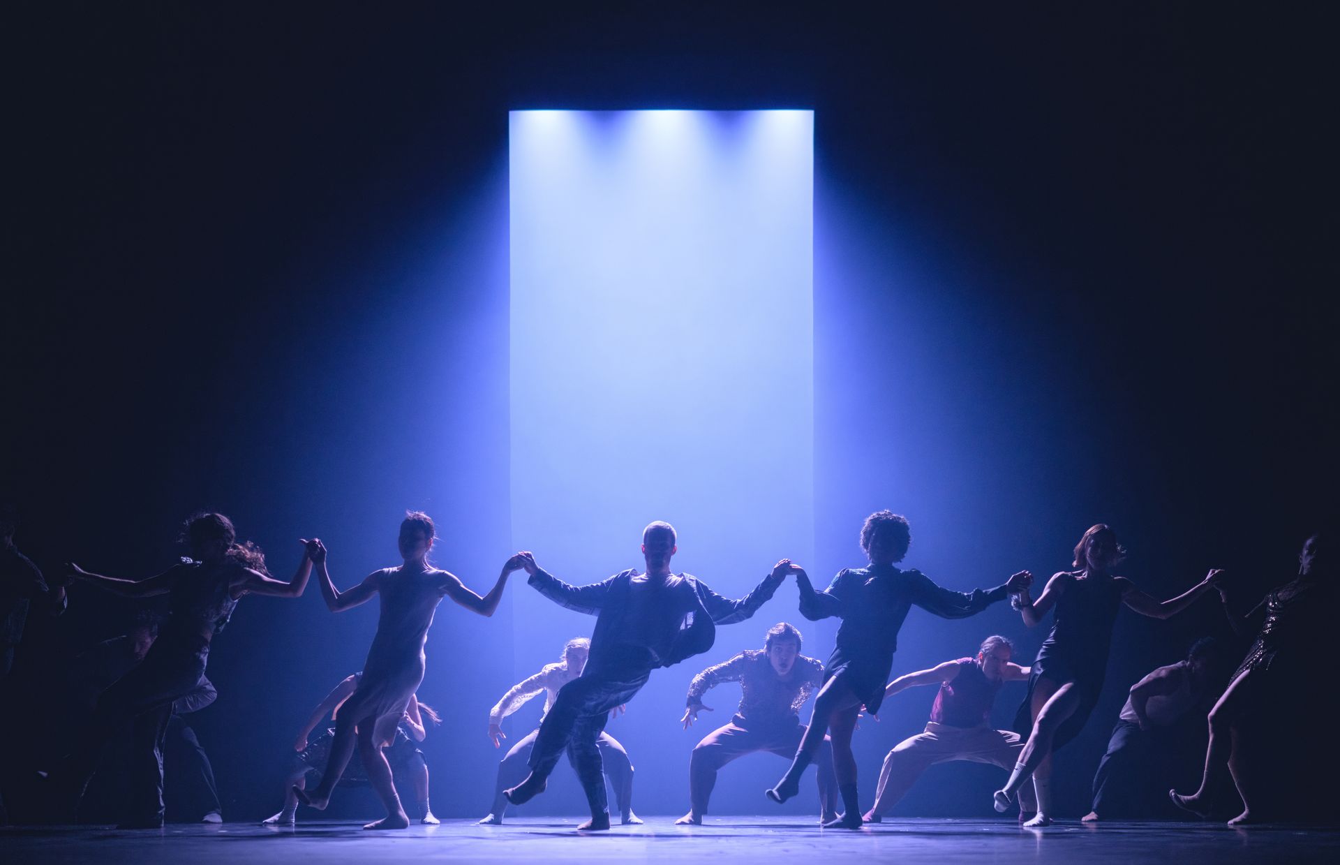 Dancers holding hands, silhouetted against a blue-lit stage. A strong spotlight shines down from above.