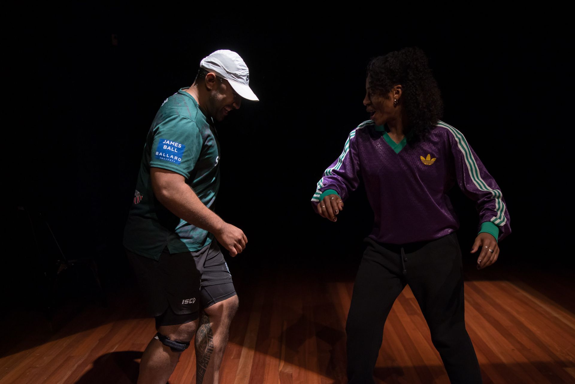 Man in green shirt and cap, woman in purple and black outfit facing each other, dancing on a wooden stage.