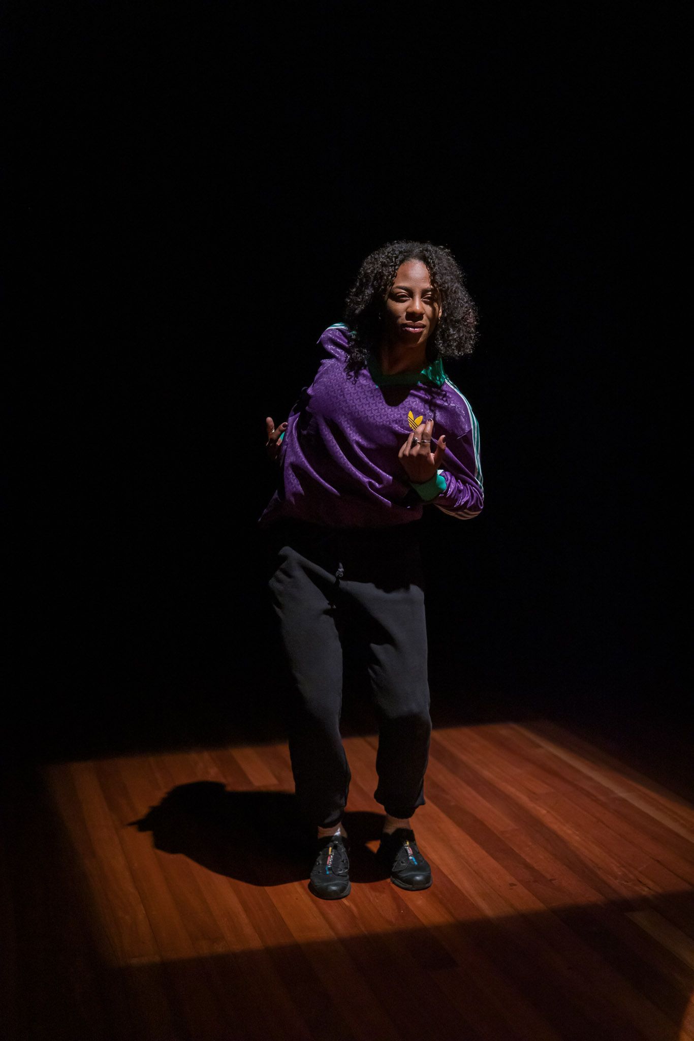 Person dancing on a wooden floor, lit by a spotlight. Wearing a purple jacket and black pants.
