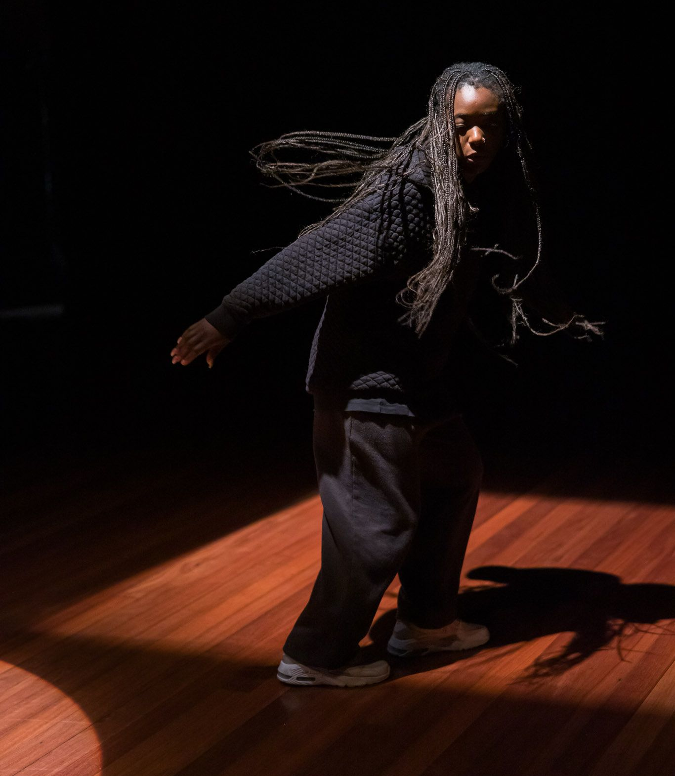 Person dancing in a spotlight on a wooden floor, wearing dark clothes, arms extended.