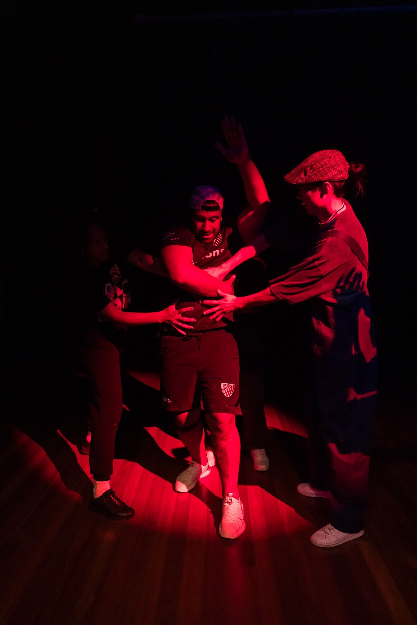Three people in a spotlight; one person is being held by the others. Red lighting. Dark background.