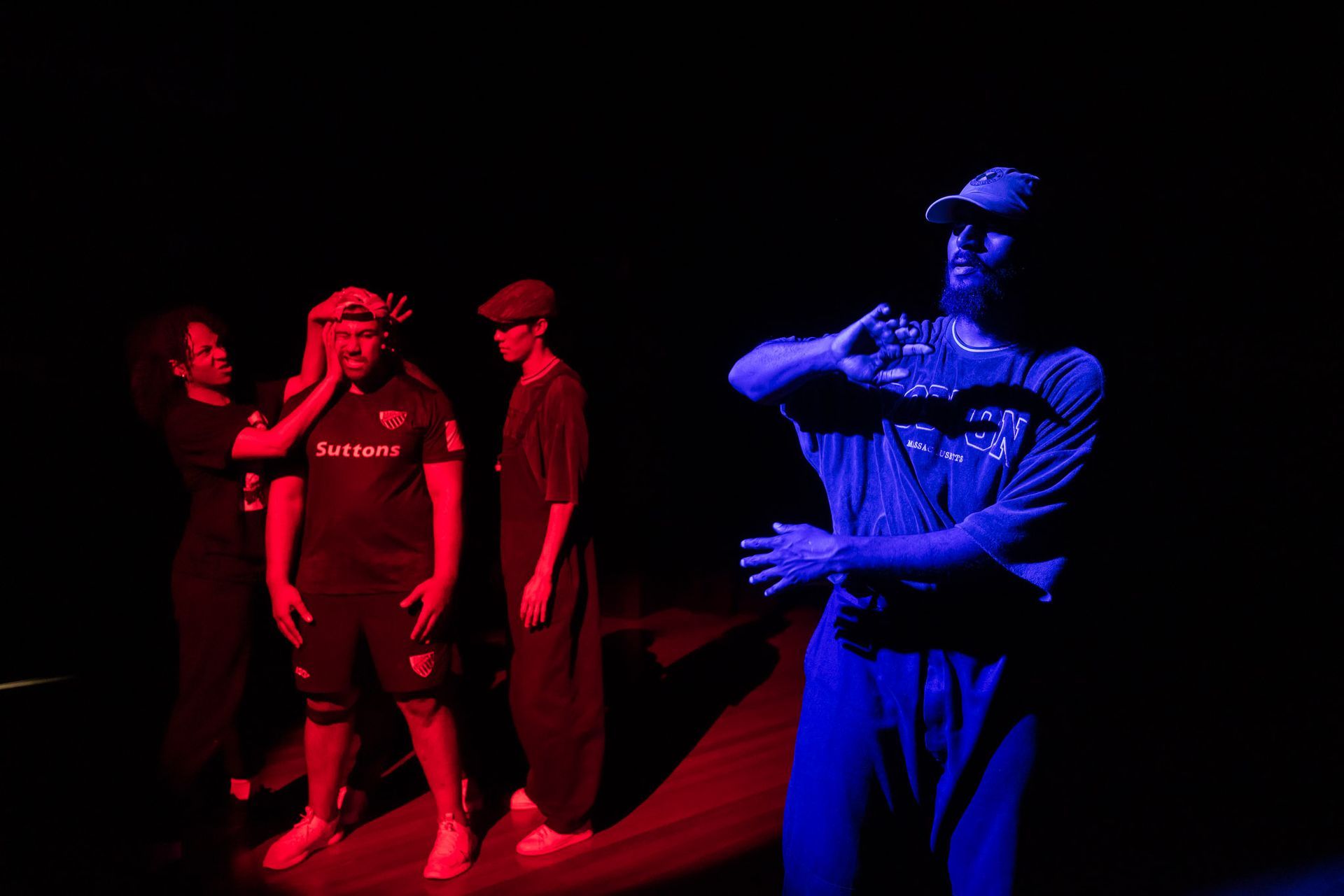 Four people on a stage. One in blue light is gesturing. Others in red light are observing. Dark background.