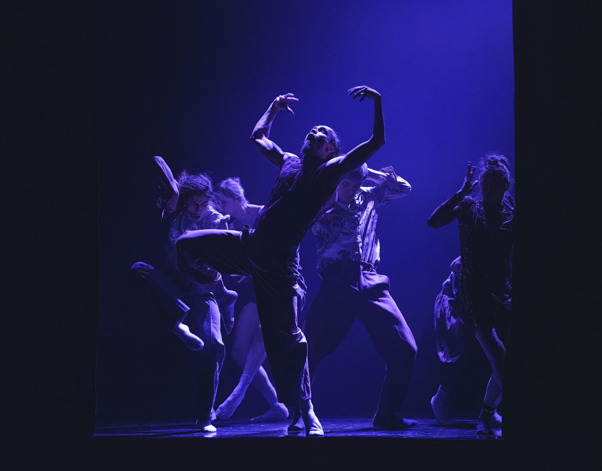 Dancers in silhouette under blue stage lighting, performing with arms raised in a group.