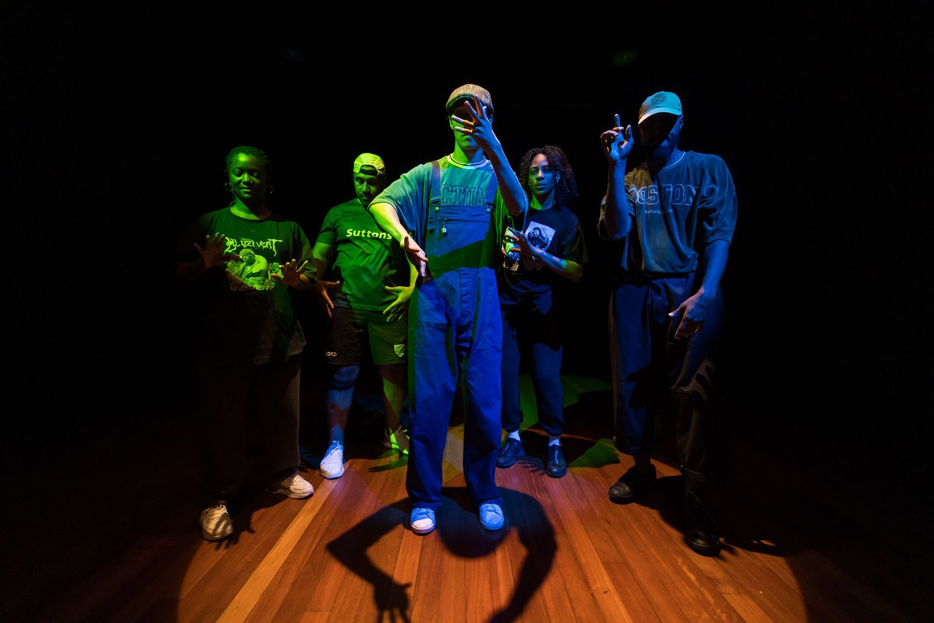 Group of six people standing on a wooden floor, lit with green and blue spotlights in a dark room.