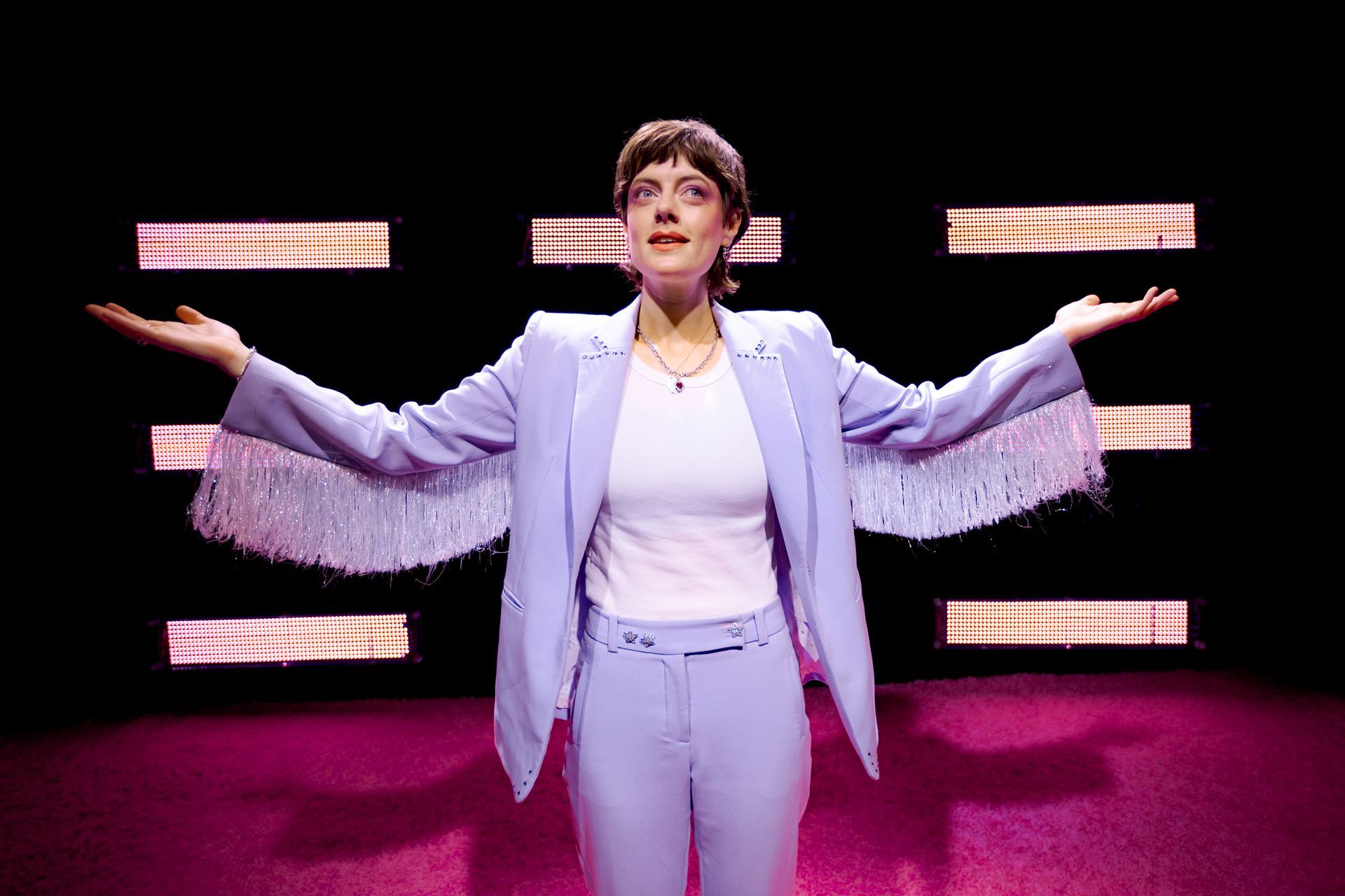 Person in lavender suit with fringed sleeves, arms outstretched, standing in front of pink lights and a black background.