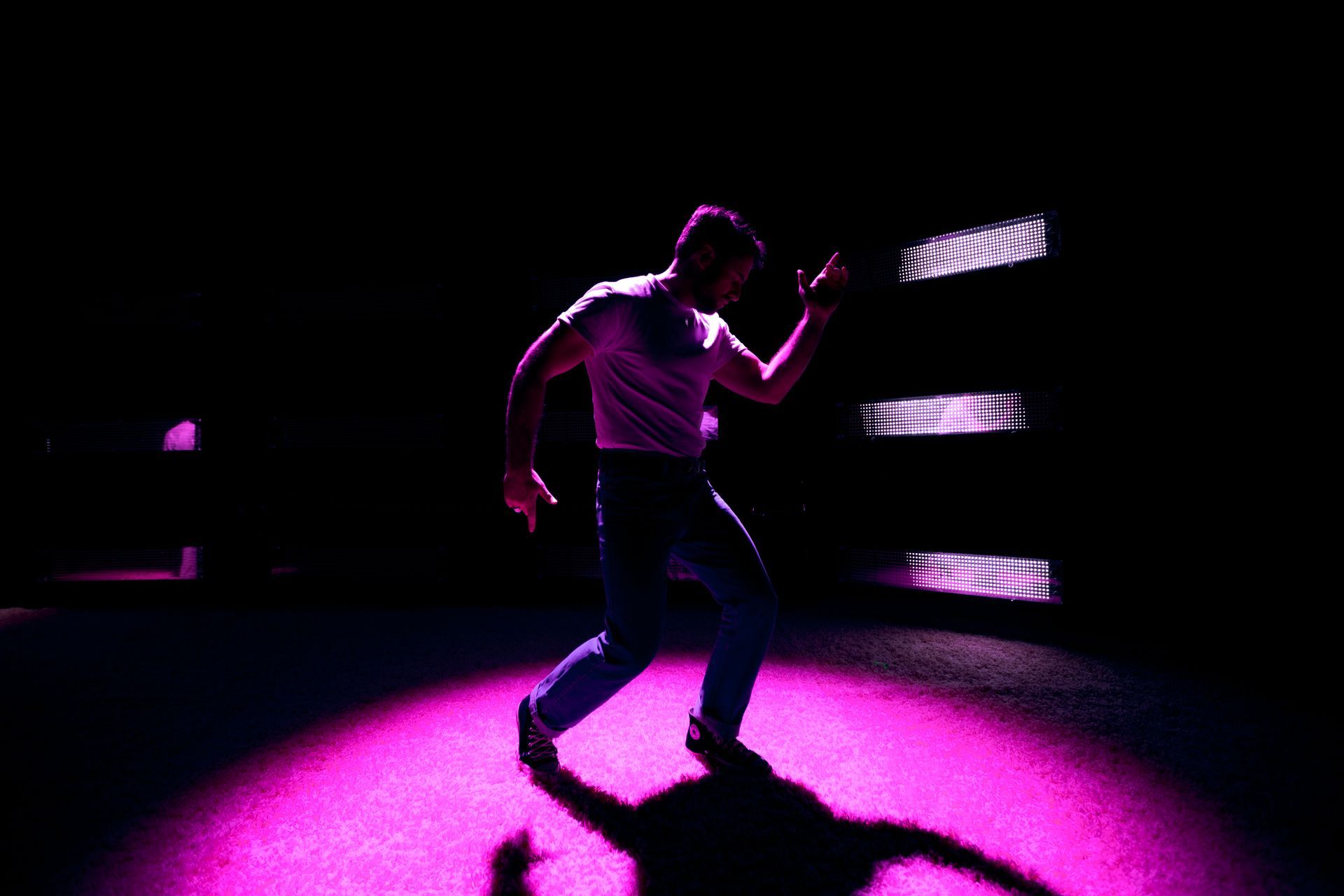Man dancing in silhouette under a pink spotlight, arms raised. Dark background with horizontal lights.