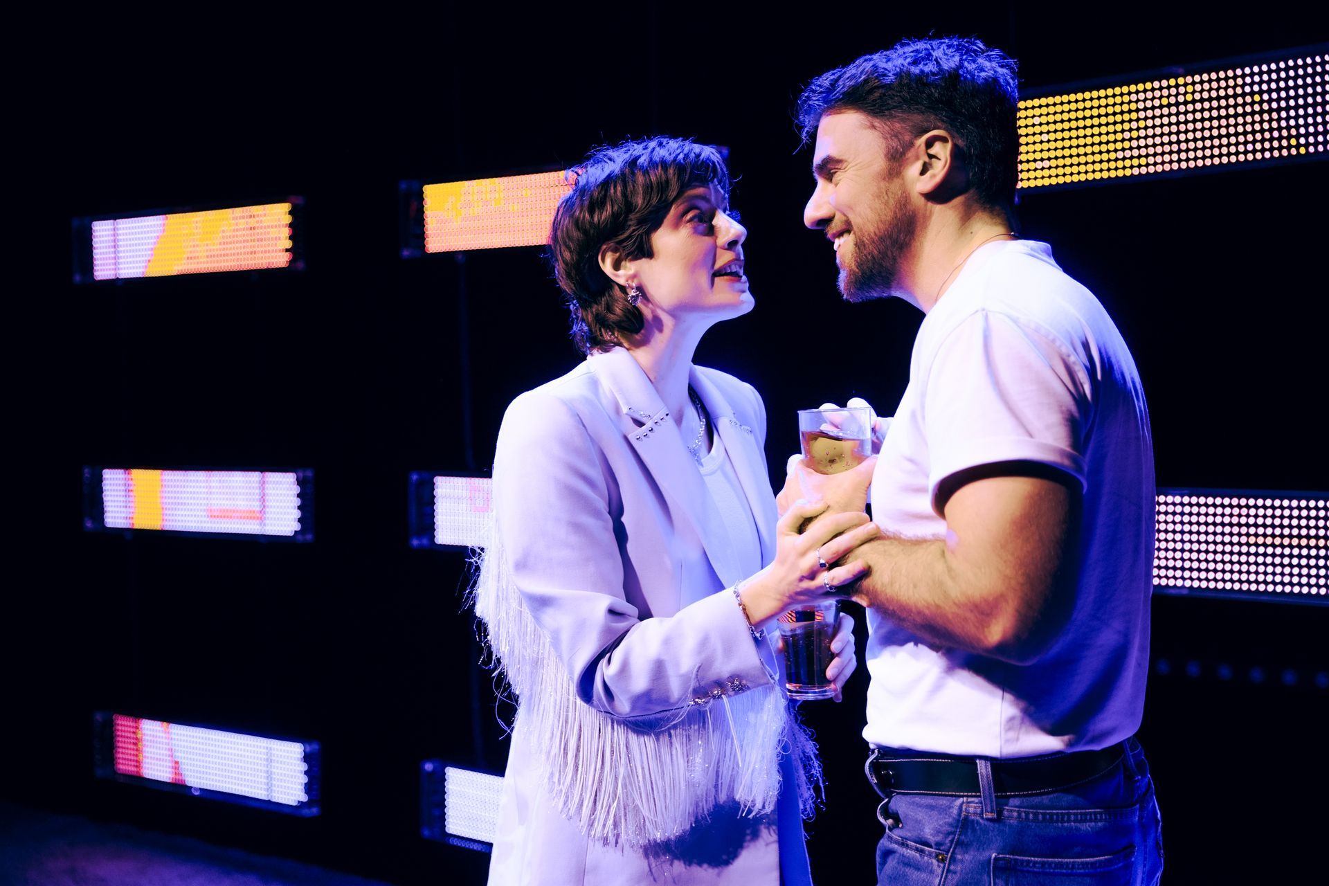 Two people facing each other, looking at each other. One in light blue suit, the other in a white shirt and jeans. Background with colorful lights.