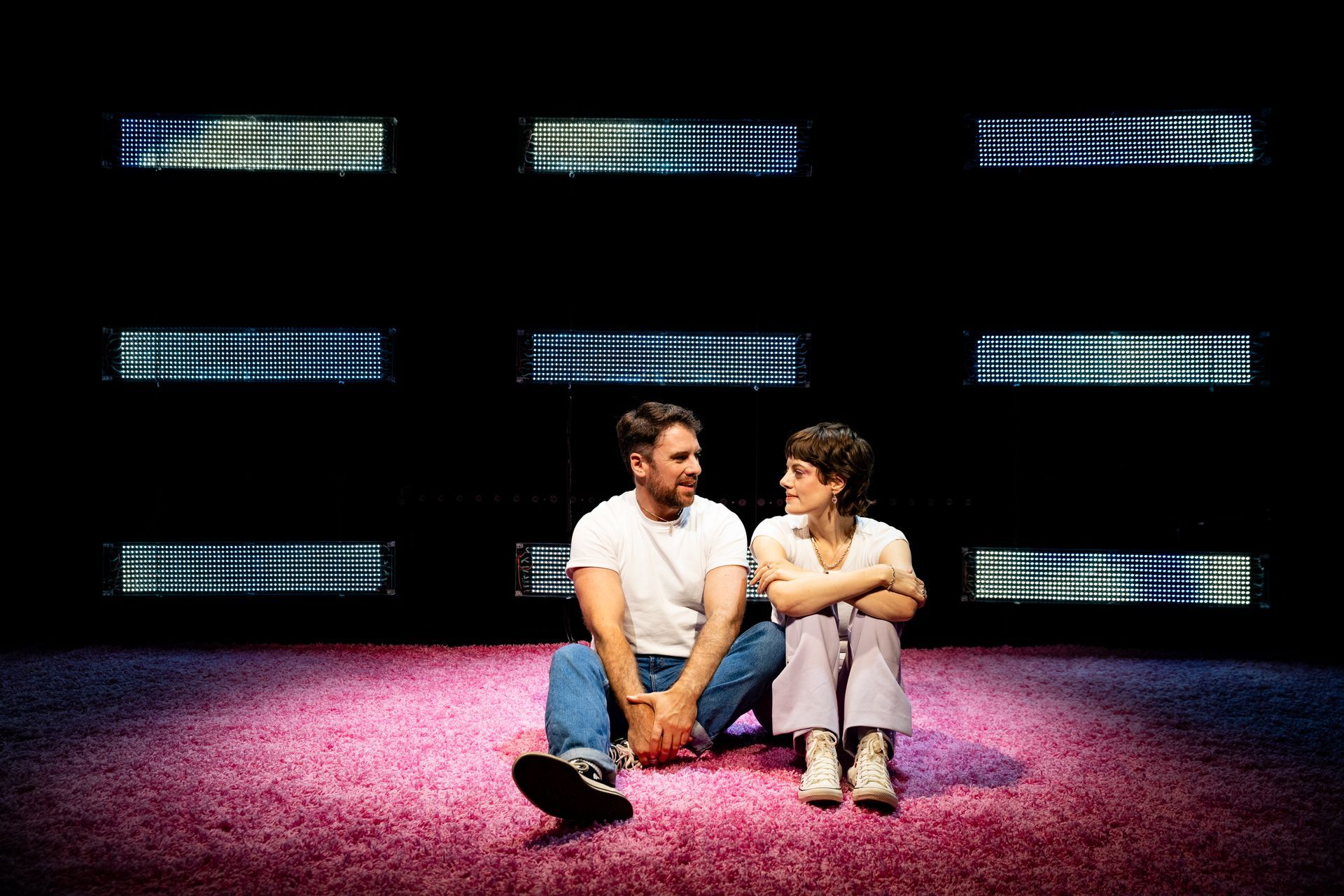 Two people sit on a pink carpet in front of a dark background with lit horizontal rectangles.