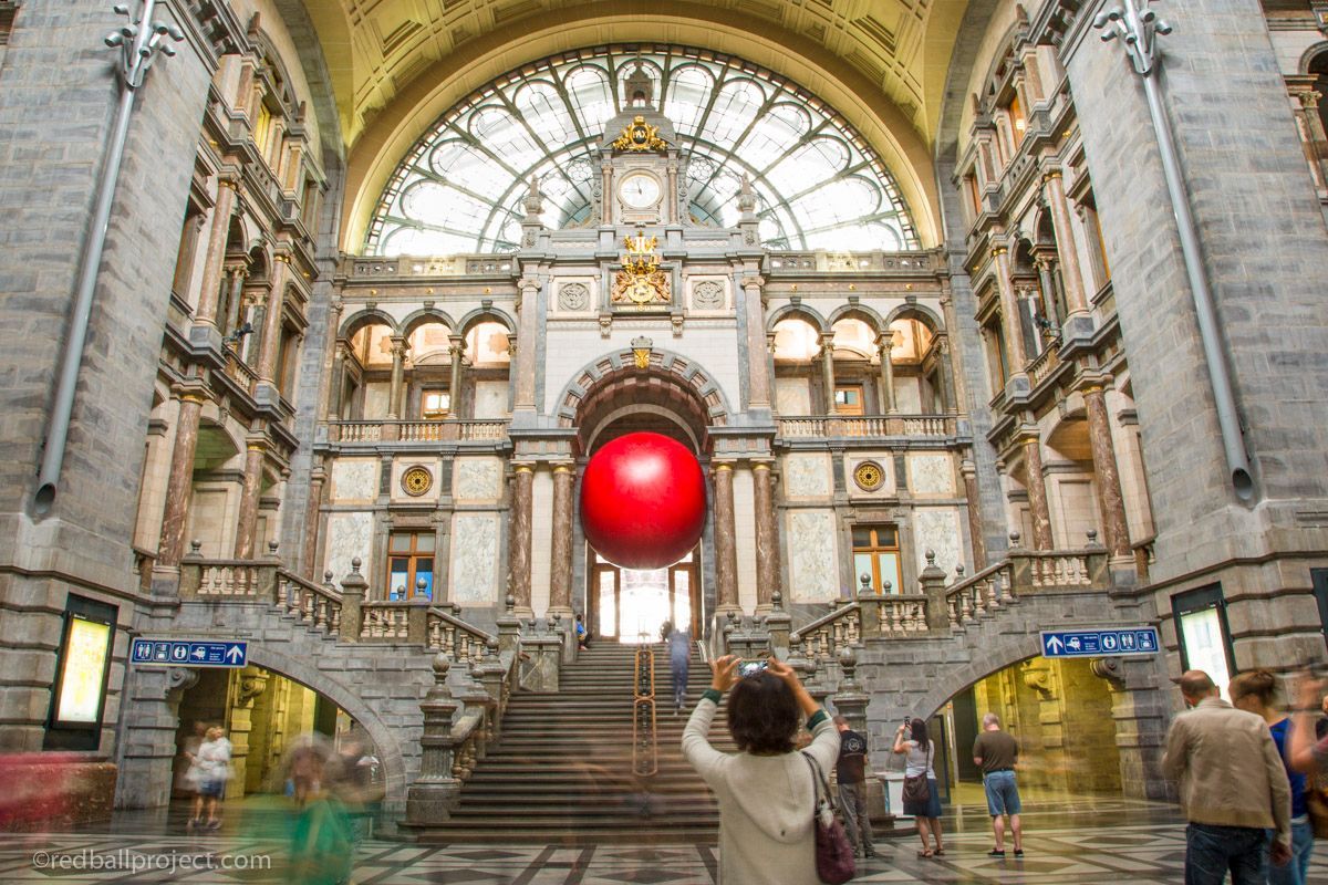 People taking pictures of a large red inflatable ball positioned i the archway of a historic building. 