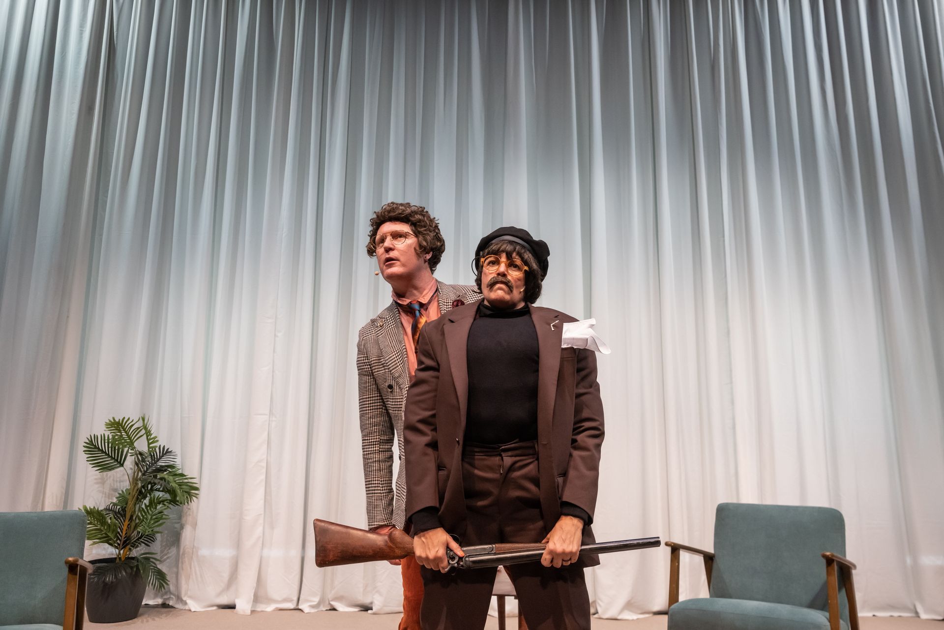 Two people on stage, one holding a rifle, the other looking concerned. White curtains, two chairs, and a potted plant.