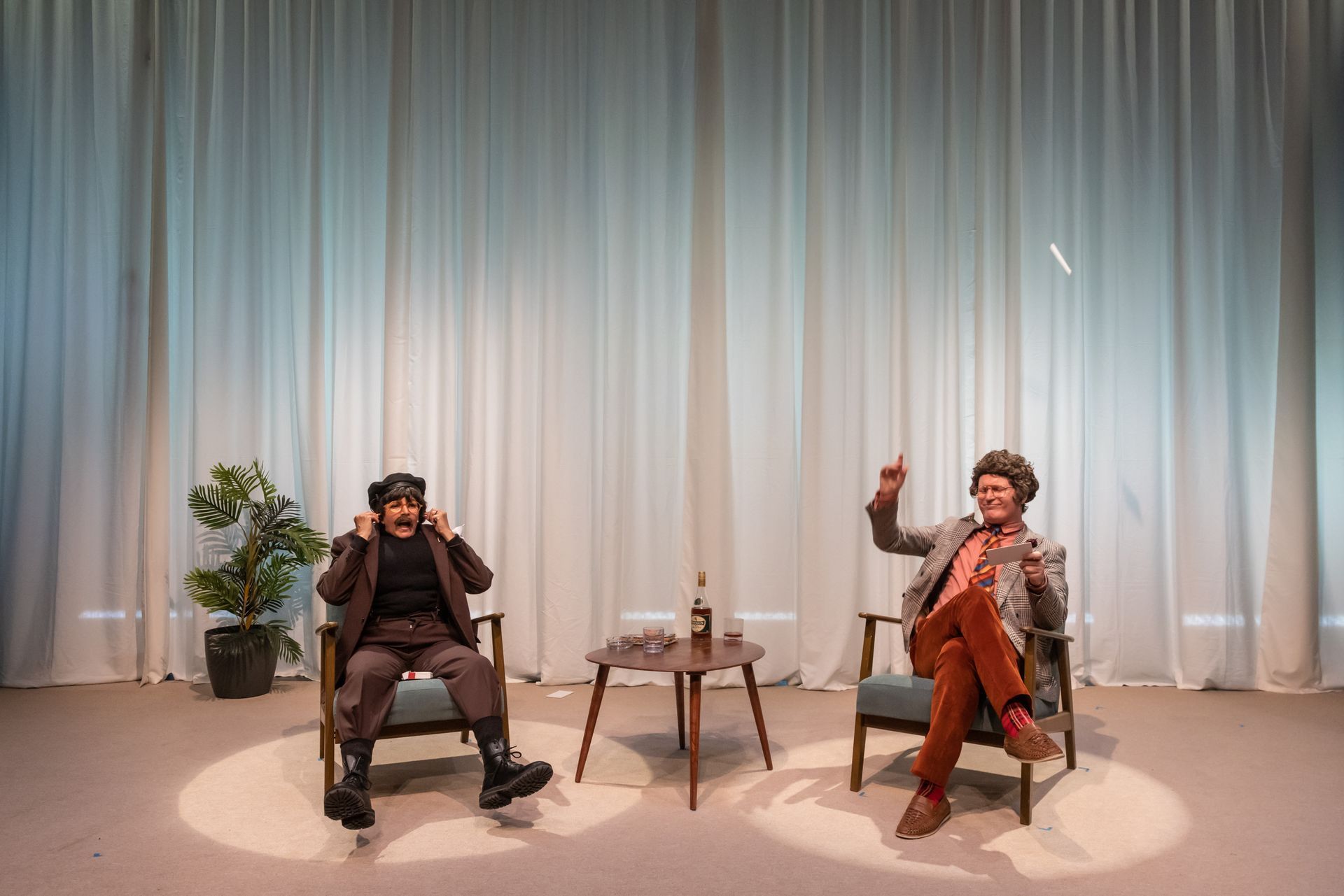 Two people in chairs on a stage. One covers ears, the other gestures. Curtains backdrop them, with a table.