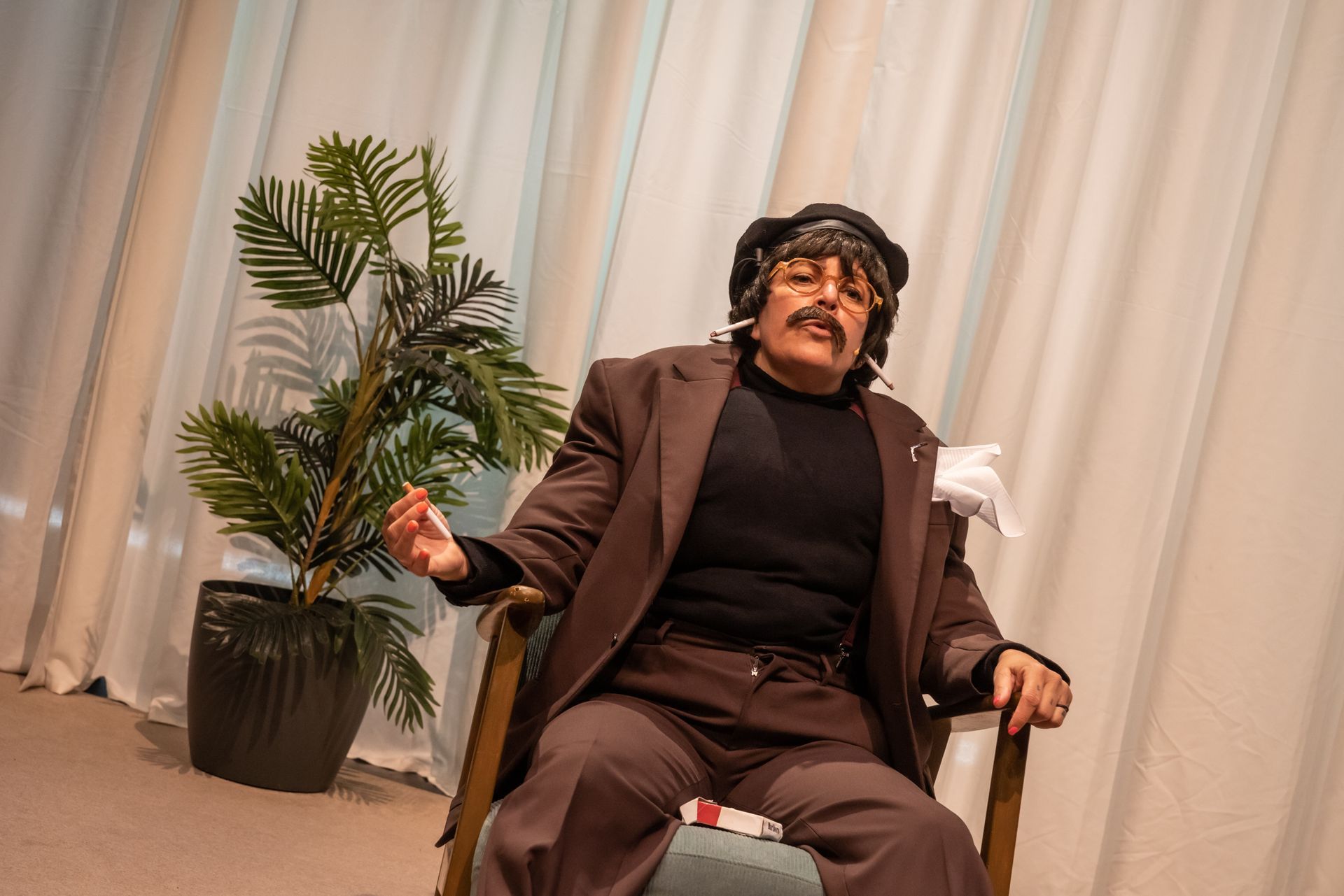 One person in a chair onstage mid-gesture. White curtains, and a potted plant.