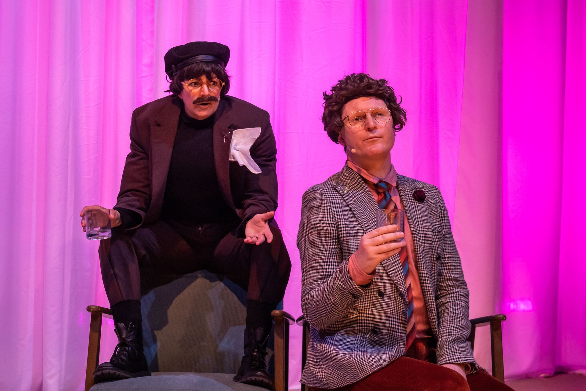 Two people in costume on a stage, one wearing a cap and dark suit, the other a zebra-striped jacket.