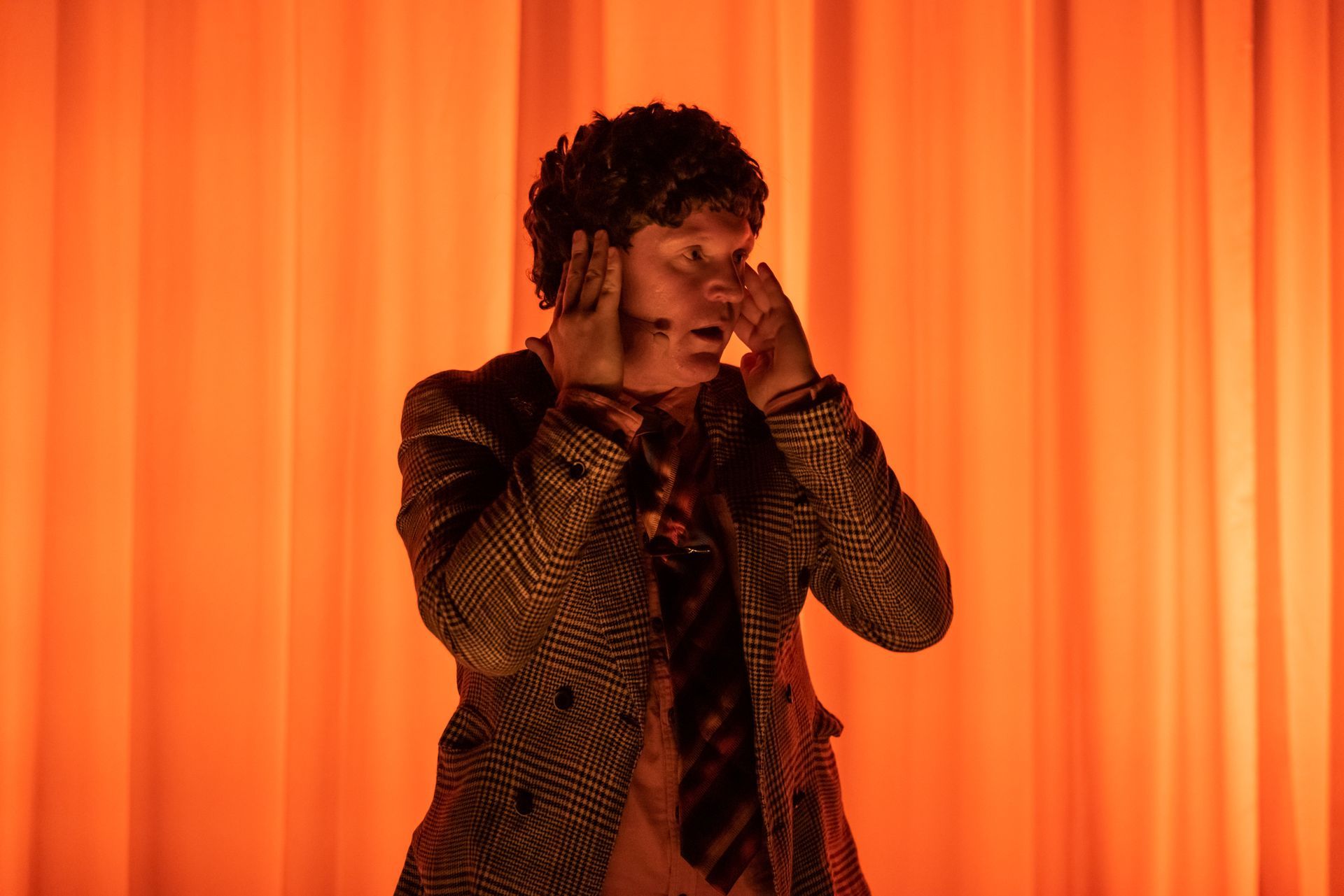Person covers ears, eyes wide, against an orange curtain backdrop. Wearing a patterned jacket, tie.