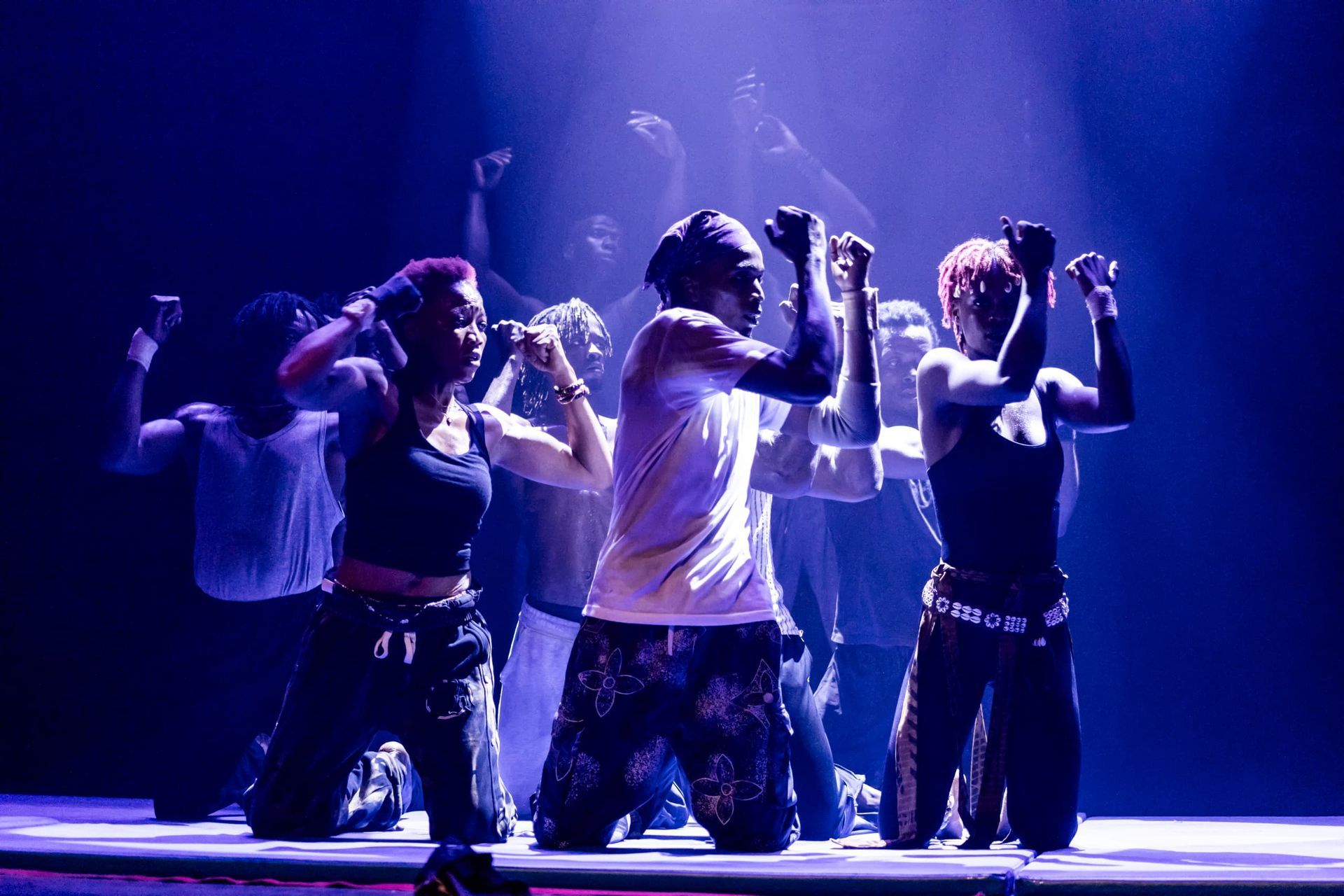 Many dancers in dramatic blue lighting gesturing with their arms up and hands in fists.