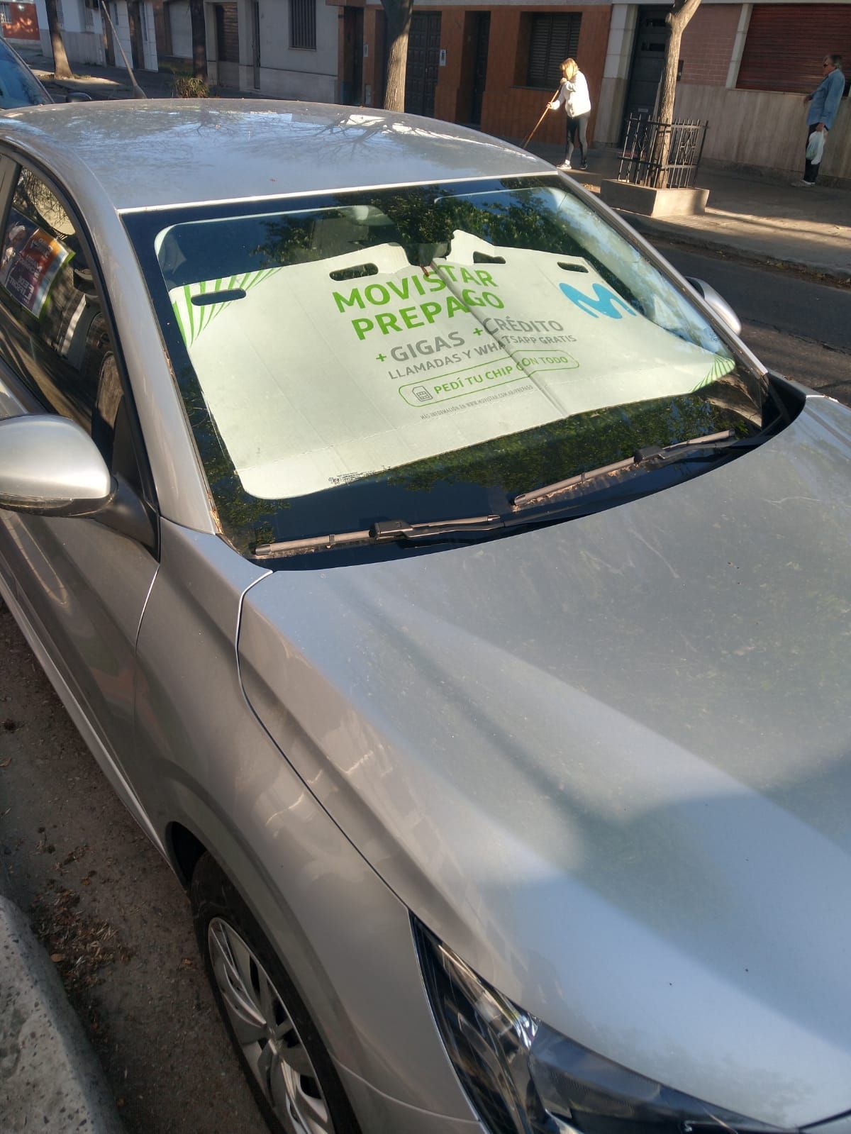 Un coche plateado con un parasol en el parabrisas está estacionado al costado de la carretera.