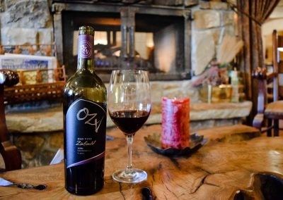 A bottle of wine and a glass of wine are on a wooden table in front of a fireplace.