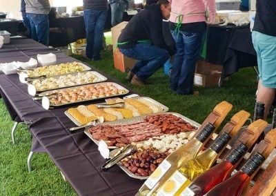 A group of people are gathered around a table filled with food and wine bottles.