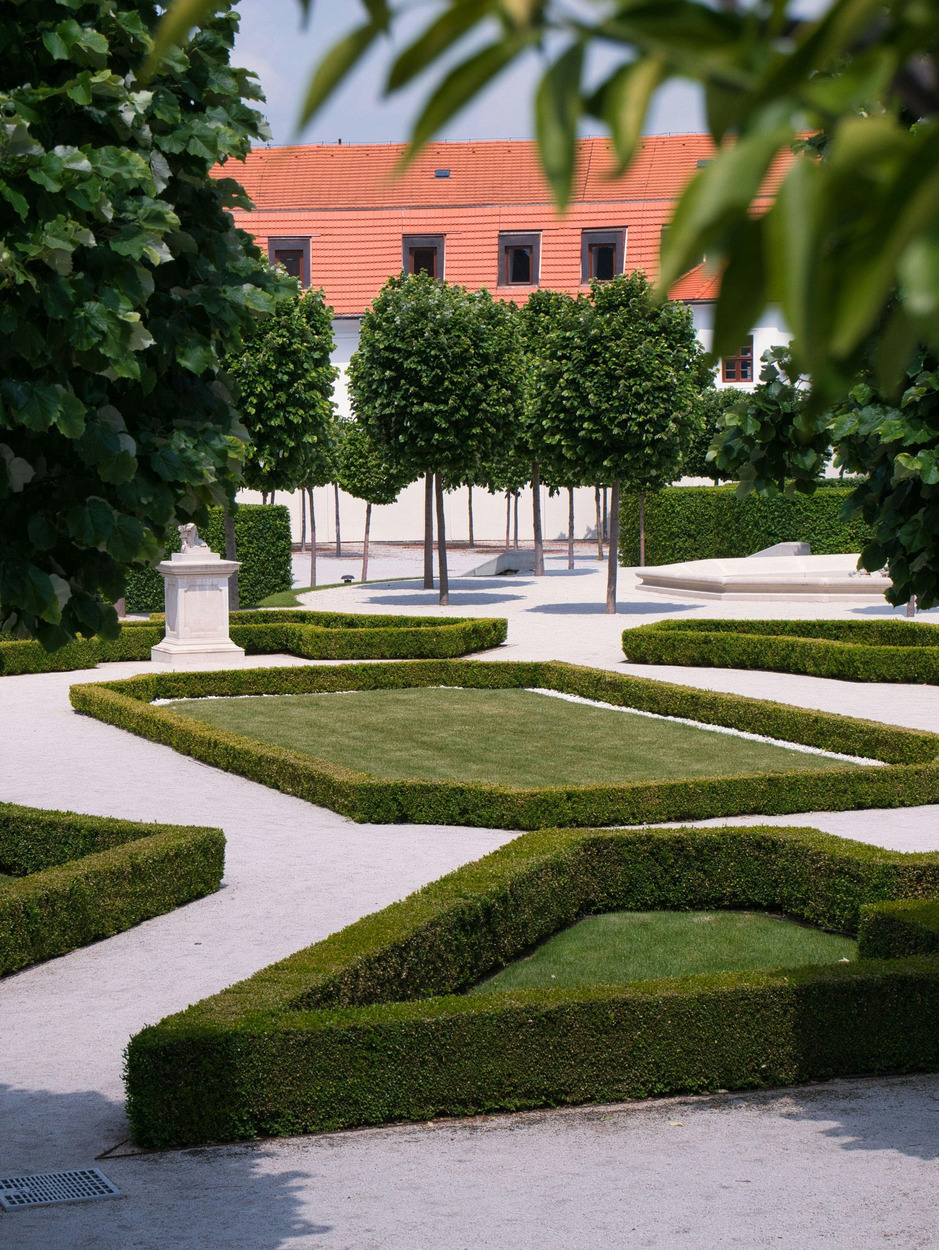 Manicured geometric hedges and gravel paths in a formal garden with trees and a red-tiled building behind