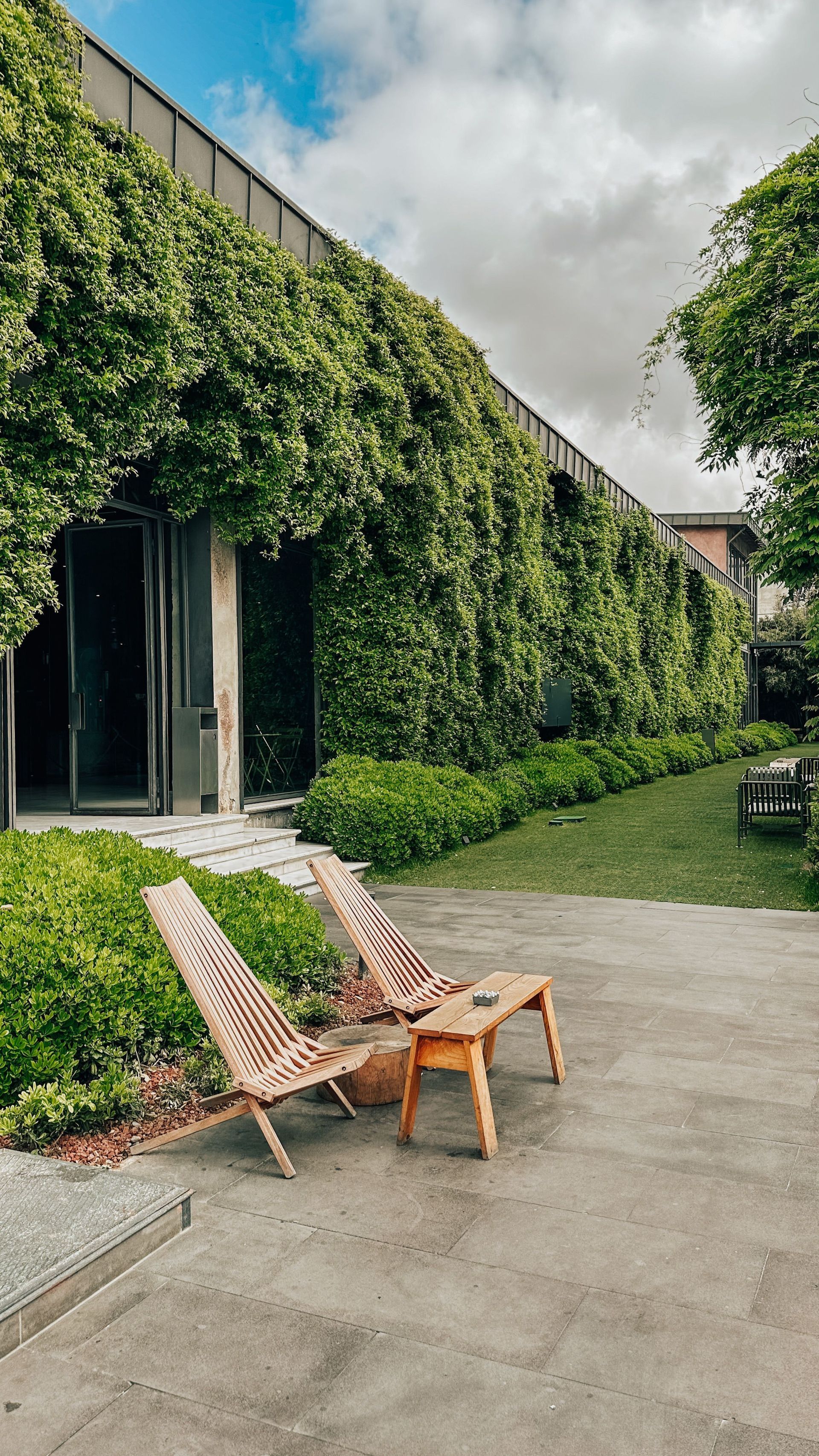 Two woven lounge chairs on a paved patio beside an ivy-covered building under a cloudy sky
