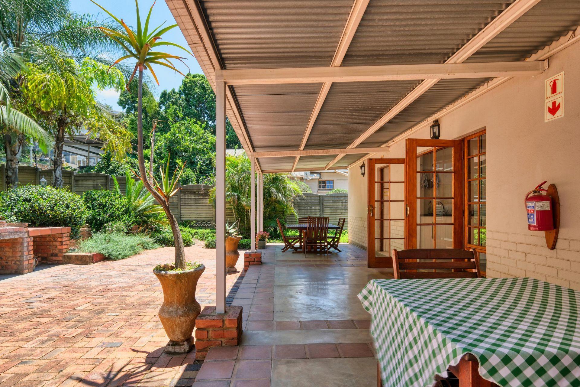 Covered patio with tiled floor, wooden chairs, potted plants, and sunlit garden outside
