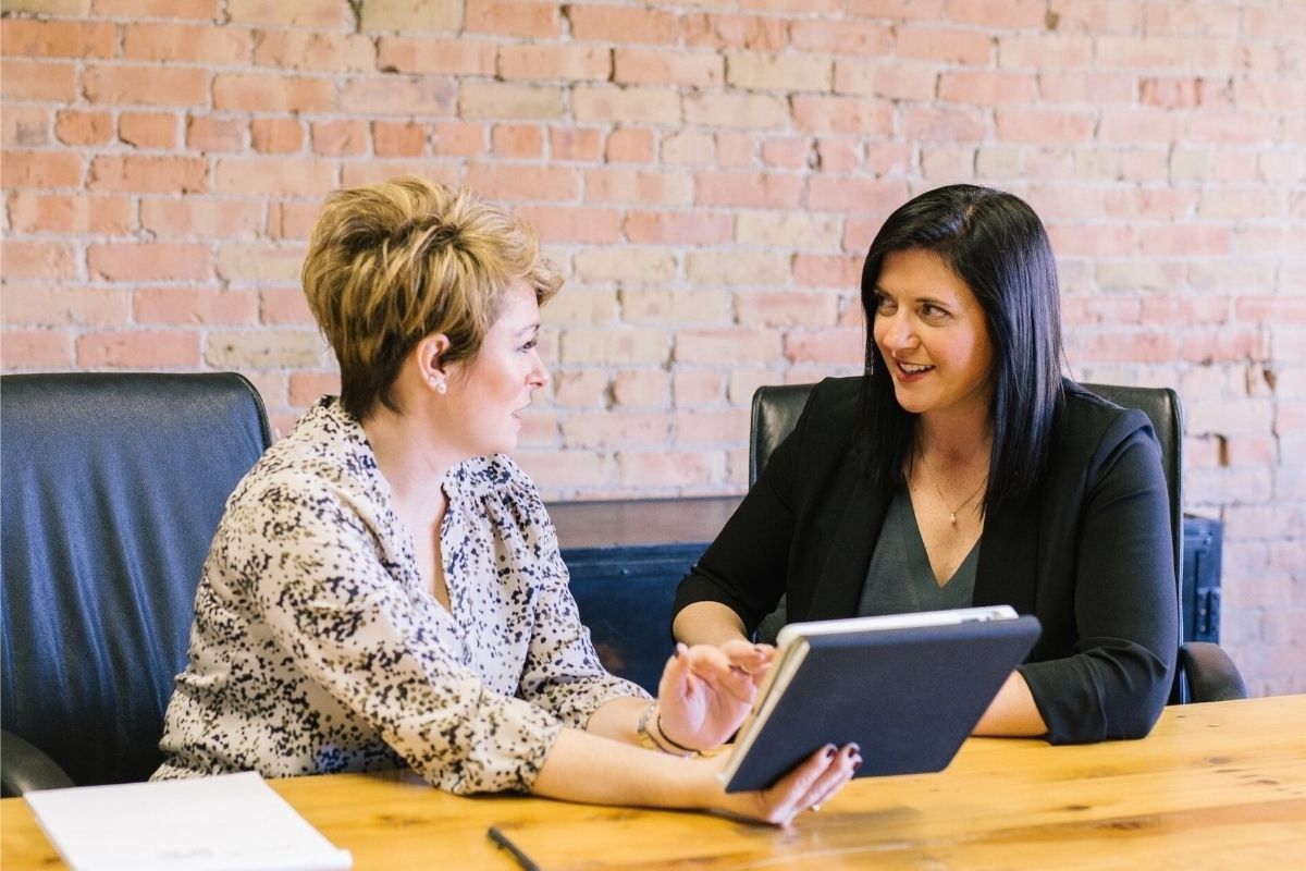 Two women are sitting at a table next to each other in front of a tablet, engaged in conversation.