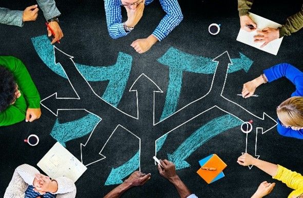 A group of people are sitting around a table with arrows drawn on a blackboard.