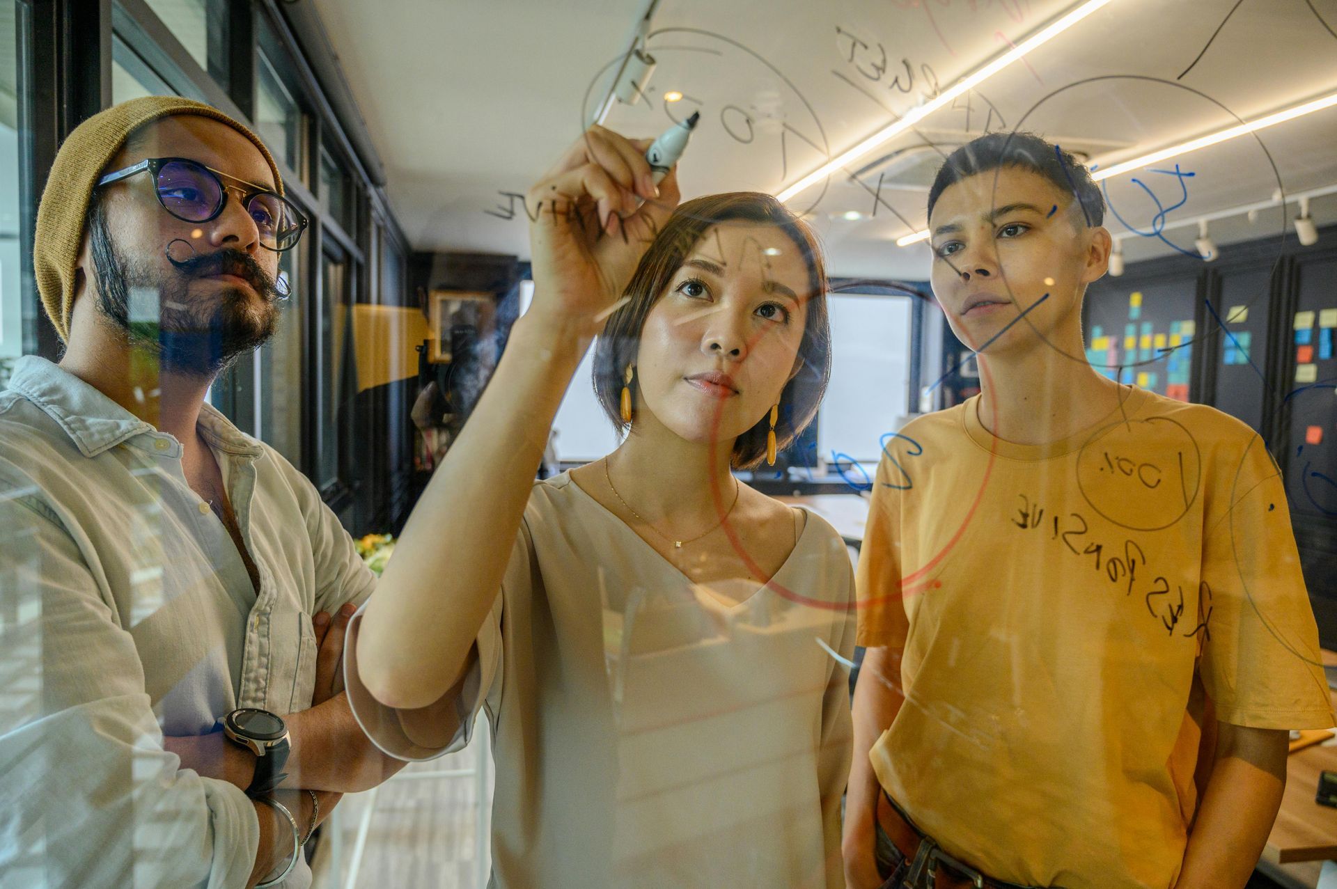 A woman is writing on a glass board while a man and a woman look on.