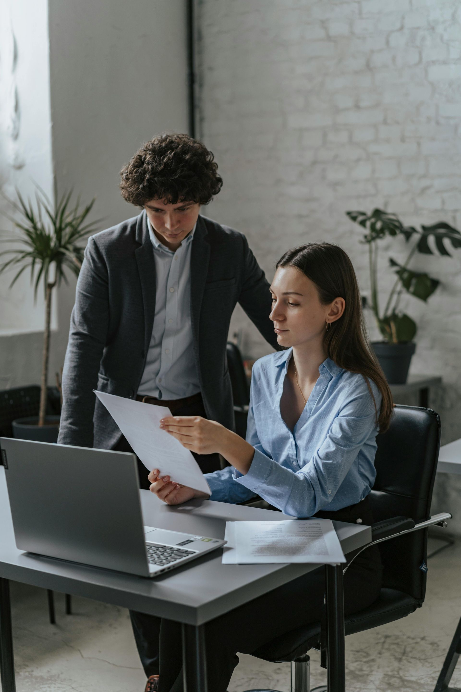 A man and a woman are looking at a piece of paper in front of a laptop computer.