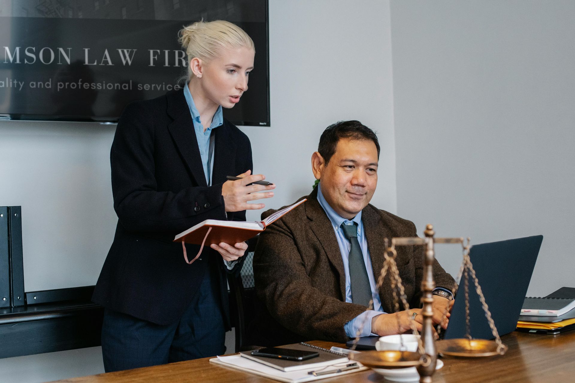 A man and a woman are sitting at a table with scales of justice and a laptop.