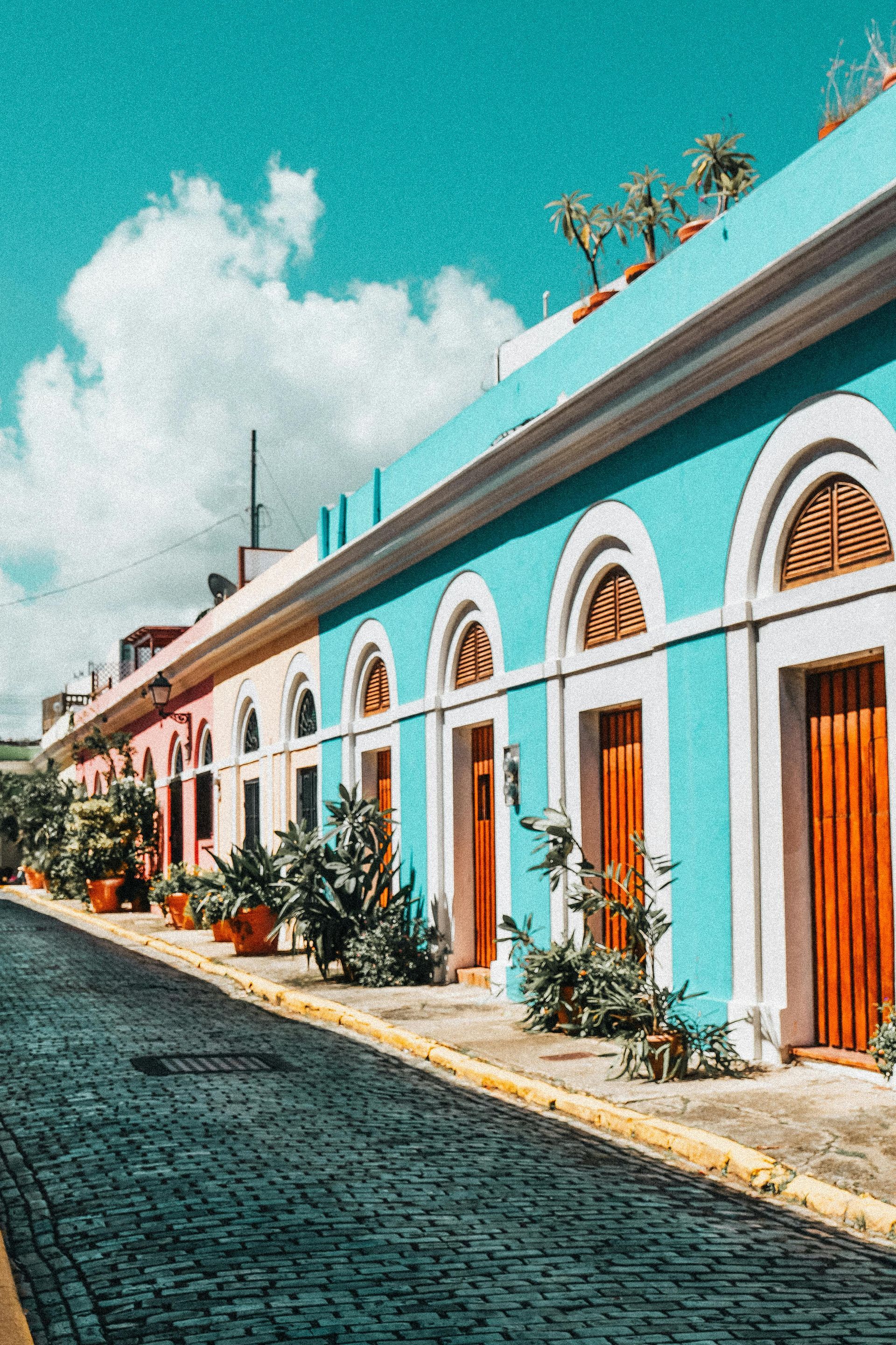 A row of pastel-colored buildings with arched doorways lines a cobblestone street under a bright, cloudy blue sky.