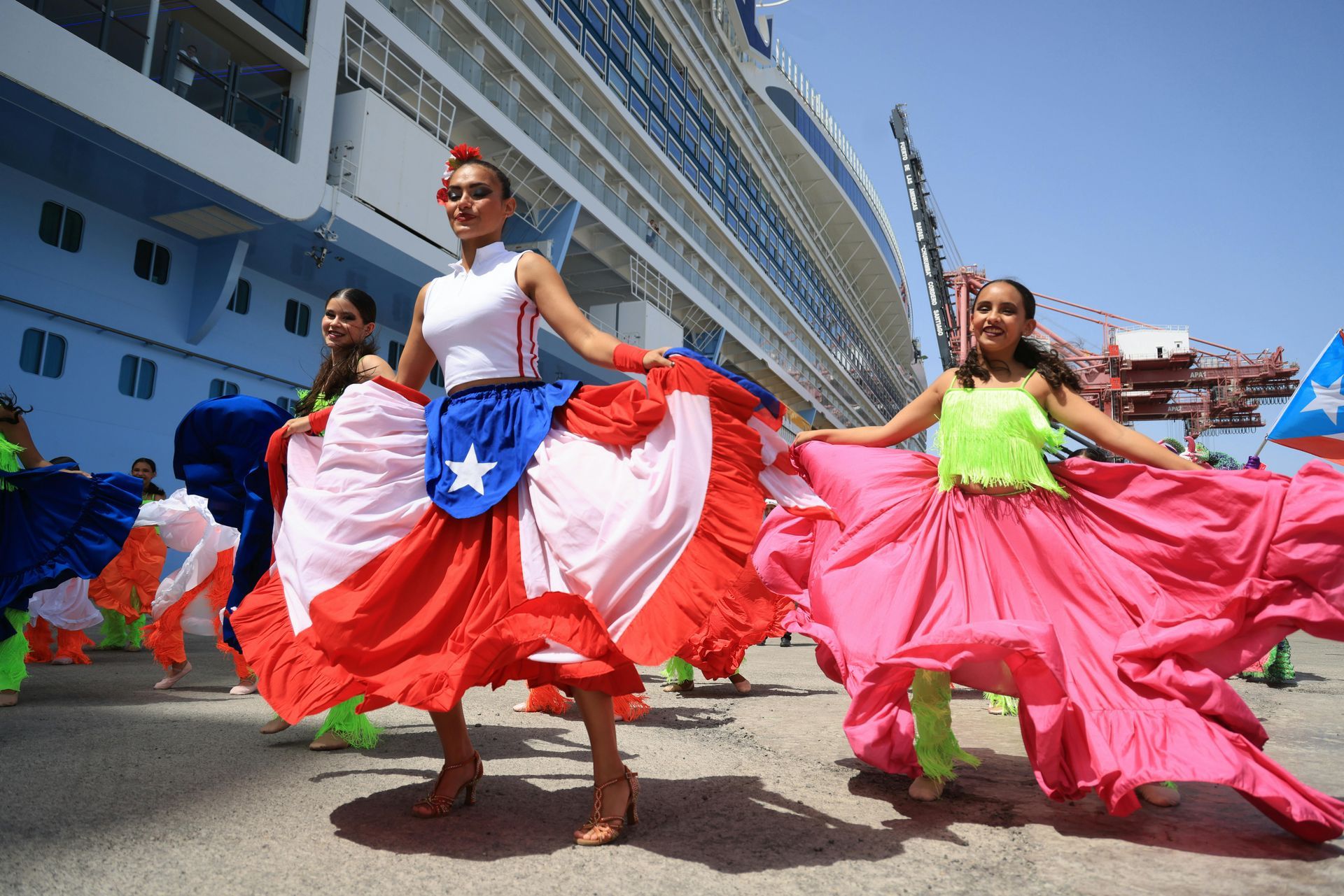 Dancers in vibrant, flowing skirts perform on a dock in front of a large cruise ship under a clear blue sky.