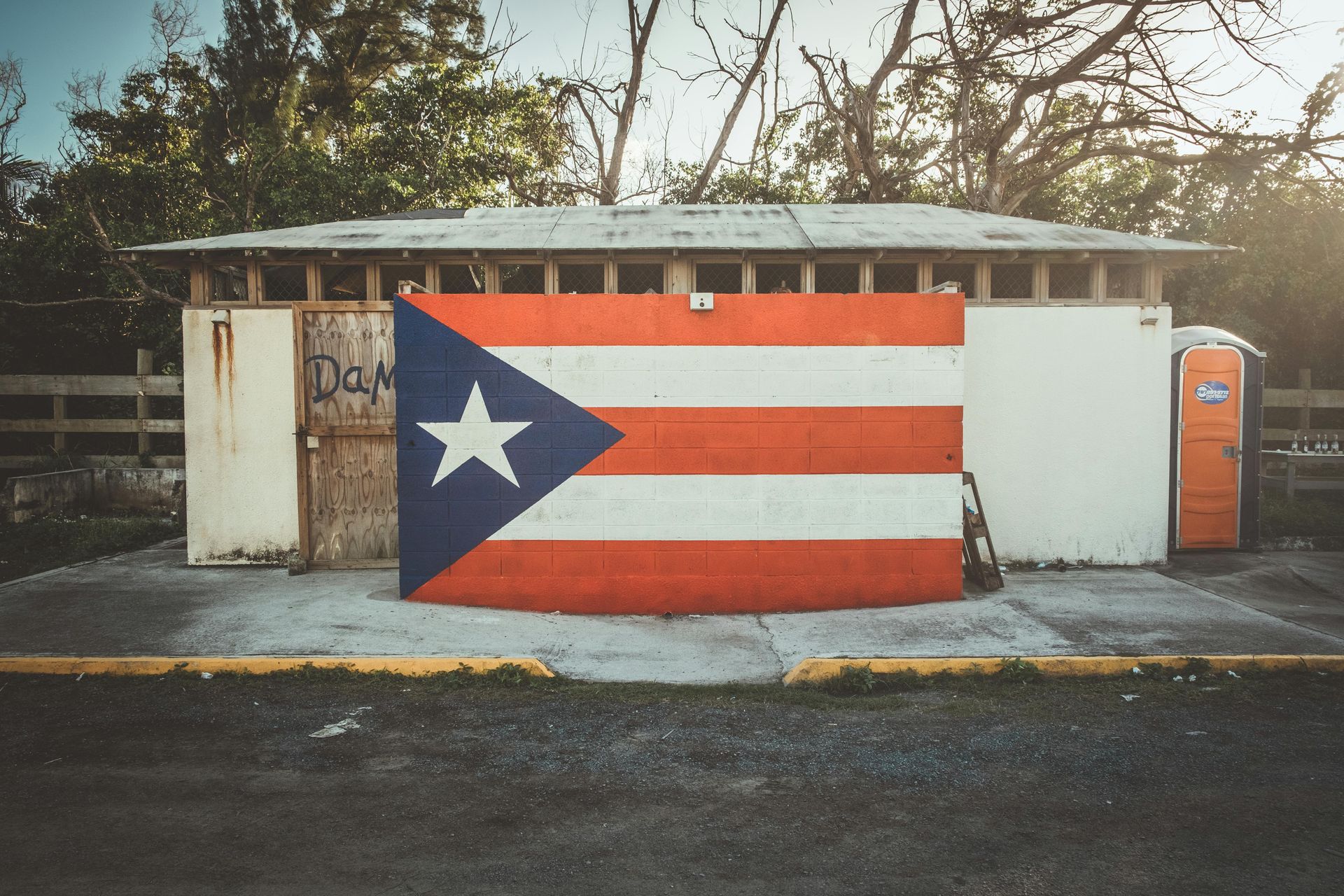 A Puerto Rican flag painted on the wall of a building exterior with an orange portable toilet nearby.
