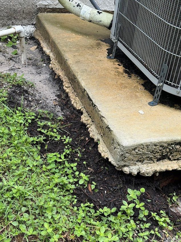 Concrete pad supporting an AC unit with overgrown weeds around the base; gray and green tones.