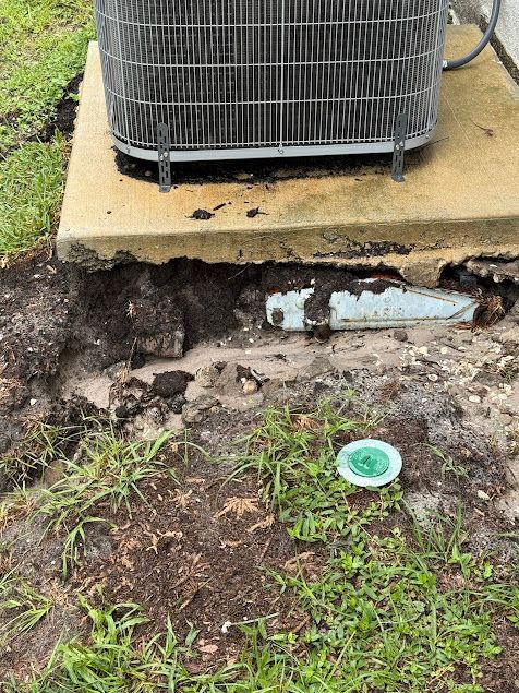 Air conditioner unit on concrete pad with exposed plumbing and ground erosion.