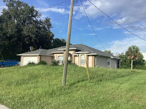 A beige house sits on a grassy hill, with power lines and a palm tree in the background.