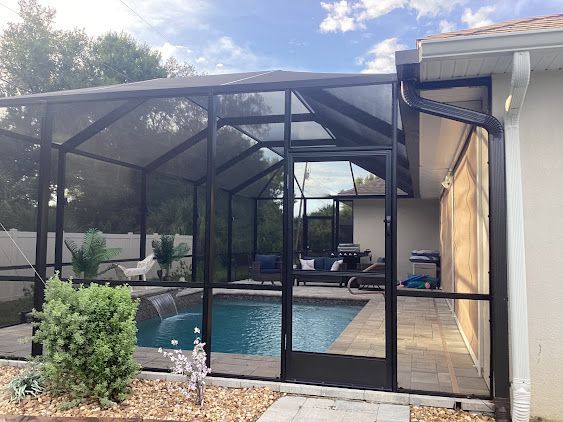 Screened-in pool enclosure with black frames, a pool, and a patio next to a house with a white gutter.