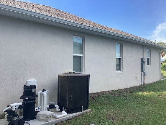 Side view of a light-colored house with windows, a pool pump, and a grassy yard under a blue sky.